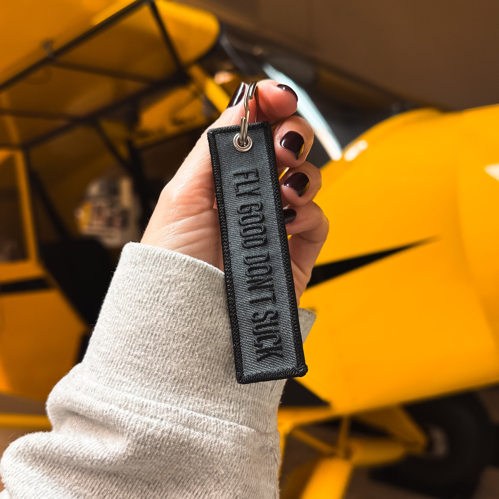 A grey and black keychain with the phrase "Fly Good Don't Suck" written on it white it is being held in front of a Super Cup airplane in a hangar