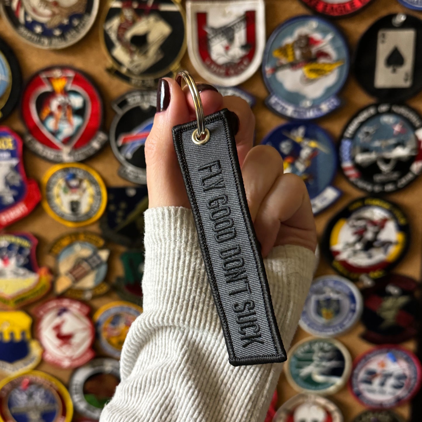 A grey and black keychain with the phrase "Fly Good Don't Suck" written on it white it is being held in front of a board full of aviation related military patches